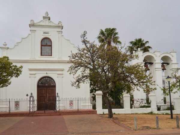 A white Cape Dutch church with a curved gable and a separate bell tower under a blue sky.